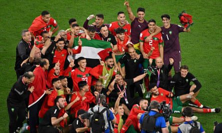 Um grupo de jogadores de camisa vermelha se reúne para uma foto em campo, gesticulando em comemoração.  Vários jogadores levantam a bandeira da Palestina no meio do grupo